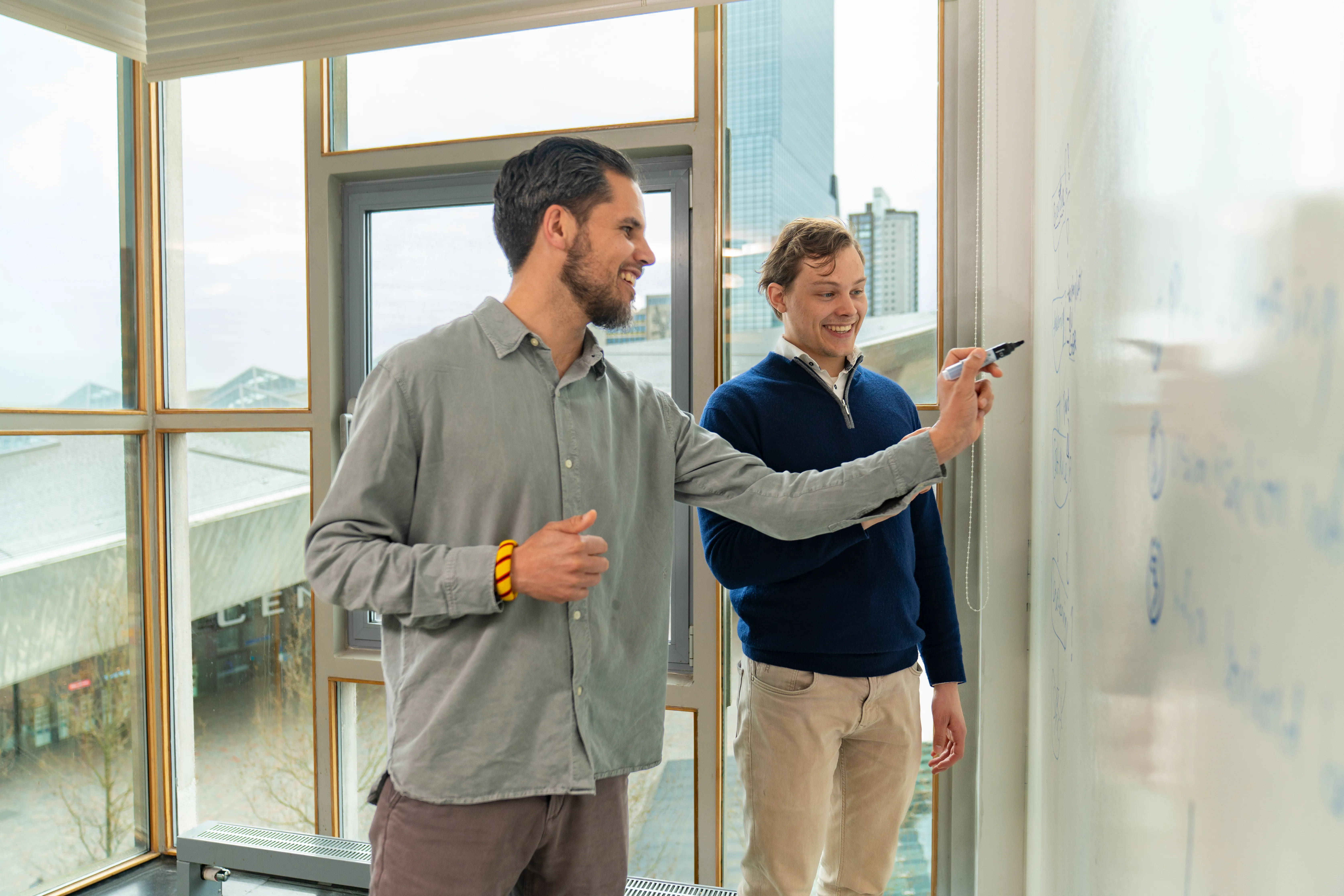 Twee collega's werken samen bij een whiteboard met uitzicht op Rotterdam